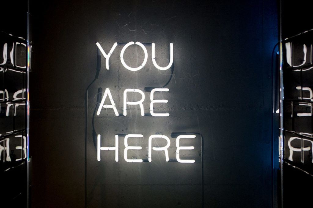White neon sign reading 'YOU ARE HERE' on a dark concrete wall, with reflections at the sides